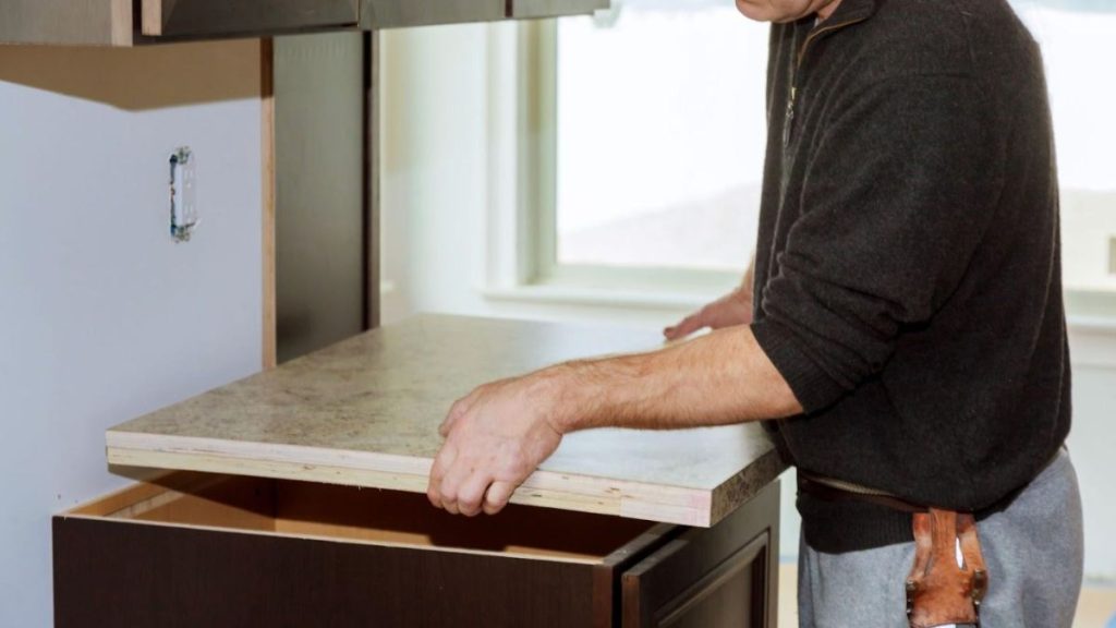 A man is installing a kitchen countertop, carefully placing a stone slab onto a cabinet base.