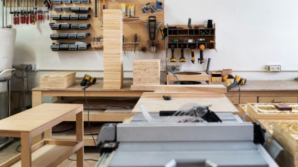 Image of a professional wood shop with organized wooden planks, power tools on the wall, and a table saw in the foreground, set up for precise woodworking projects.